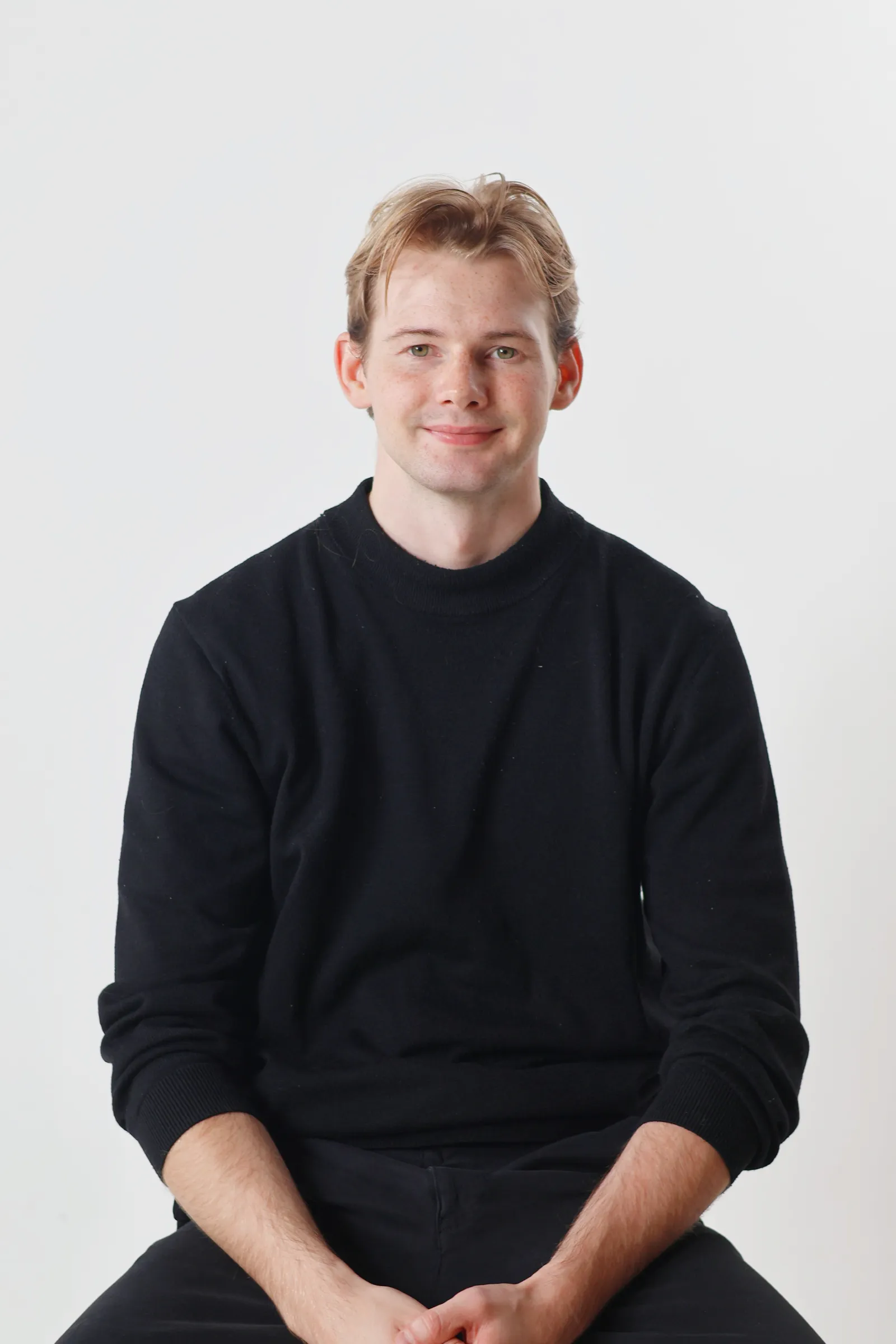 A portrait of a young man with blond hair and a black sweater sitting in front of a white background.